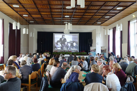 Menschen sitzen an Tischen in der Aula des Campus in Bockhorst, im Hintergrund ist ein historisches Foto einer Unterrichtssituation auf einer Leinwand zu sehen.